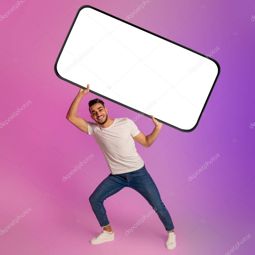 Cheerful young Arab man holding huge heavy smartphone with empty screen in neon light. Mockup