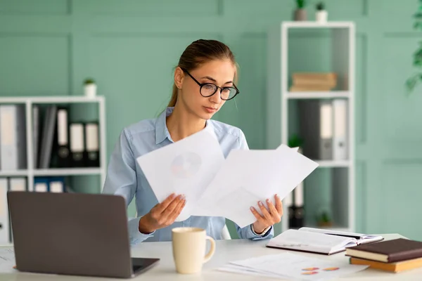 Geconcentreerde zakenvrouw werkend met rapporten en documenten, zittend met laptop op de werkplek in kantoorinterieur — Stockfoto