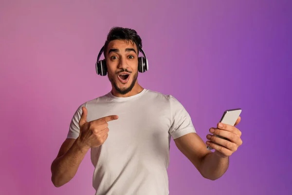 Förvånad ung arabisk kille i hörlurar pekar på smartphone, chockad över online-försäljning i neon ljus — Stockfoto