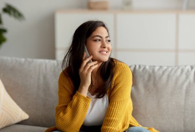 Orta Doğulu mutlu bayan akıllı telefondan konuşuyor, evdeki kanepede oturuyor ve boş yere bakıyor.