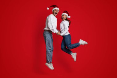 Aşırı mutlu çift Noel Baba şapkası takıyor, el ele tutuşuyor ve kırmızı arka planda zıplıyorlar.