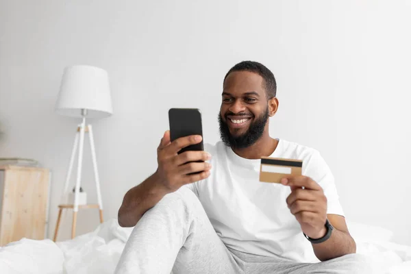 Joven afroamericano alegre con barba hace compras remotas con smartphone y tarjeta de crédito —  Fotos de Stock