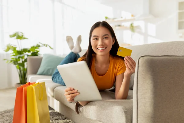 Happy korean woman lying on sofa with tablet pc and credit card, shopping online and purchasing clothes from home — Stock Photo, Image