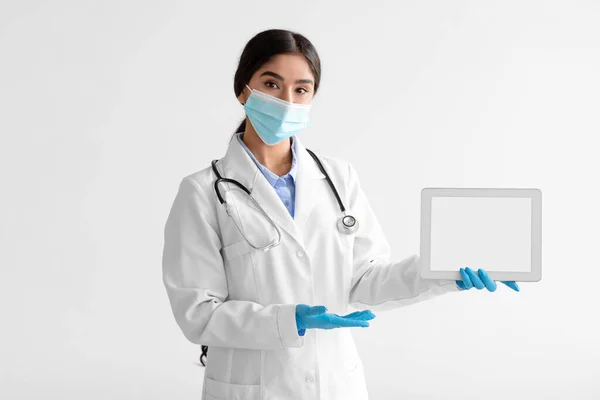 Joven médico hindú de abrigo con estetoscopio en máscara protectora, guantes sujetan la tableta, muestran la mano a la pantalla en blanco —  Fotos de Stock