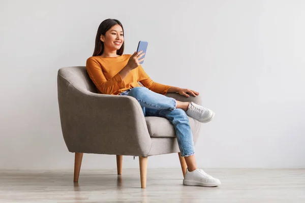 Mujer asiática usando el teléfono móvil, charlando en la web, trabajando o estudiando en línea, sentado en sillón contra la pared blanca —  Fotos de Stock