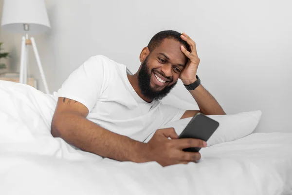 Feliz joven afroamericano masculino con barba escribiendo en el teléfono y viendo vídeo en la cama blanca en el dormitorio — Foto de Stock