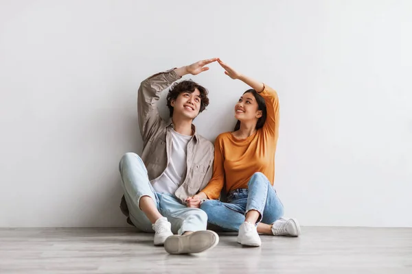 Positivo millennial asiático pareja haciendo casa techo con sus manos, sentado en el suelo en nuevo apartamento vacío — Foto de Stock