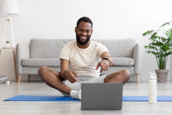 Happy young african american bearded man on floor on mat in living room interior with bottle of water — 图库照片