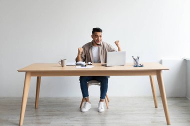 Arap adam dizüstü bilgisayar kullanarak başarıyı kutluyor. Yumruklarını sallayarak evet diyor.