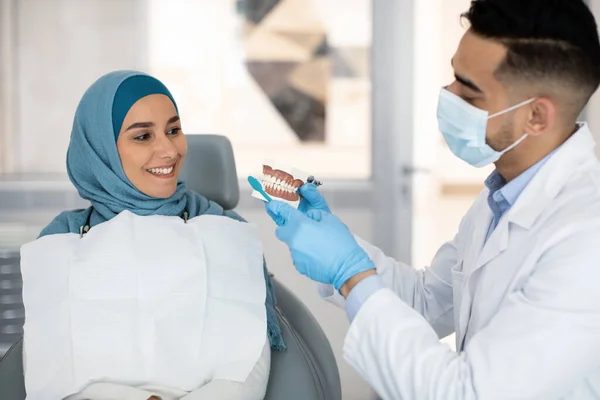 Dentista masculino segurando modelo de mandíbula, explicando higiene dentária para muçulmano paciente do sexo feminino — Fotografia de Stock