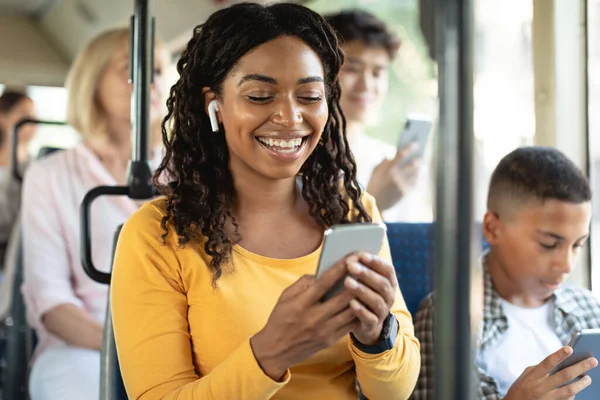 Senhora preta feliz usando smartphone usando fones de ouvido no ônibus — Fotografia de Stock