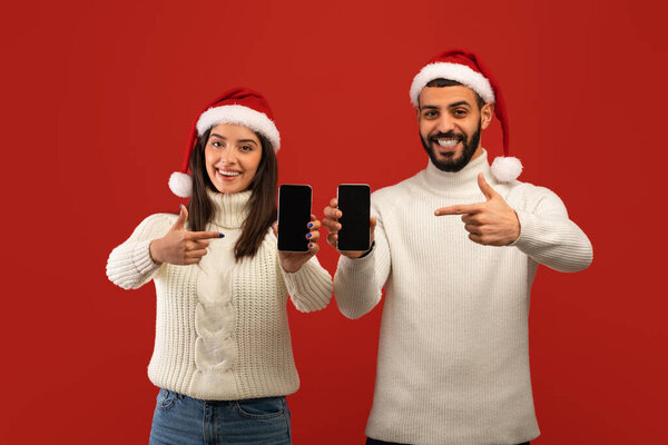 Christmas advertisement. Arab spouses pointing fingers at cellphone screens, wearing Santa hats over red background — Stock Image Christmas advertisement. Arab spouses pointing fingers at cellphone screens, wearing Santa hats over red background Royalty Free Stock Photos