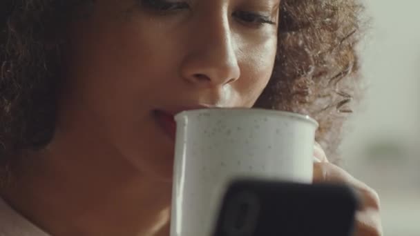 Close-up portret van onherkenbaar Afrikaans amerikaanse vrouw drinken thee of koffie en web surfen online op mobiele telefoon — Stockvideo