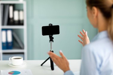 Genç iş blogcusu cep telefonuyla video kaydediyor, cep telefonuyla konuşuyor ve tripodda boş ekran var.