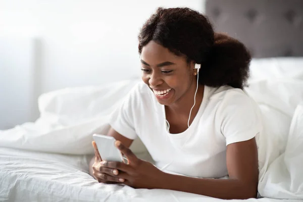 Glückliche junge schwarze Frau liegt mit Smartphone im Bett — Stockfoto