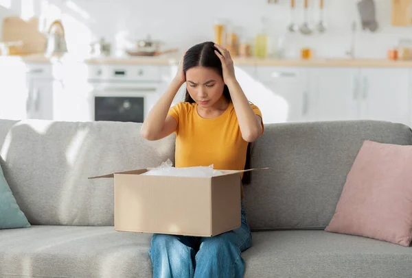 Slechte bezorgservice. Ontevreden aziatische dame zit op de bank met kartonnen doos, ontvangen pakket met verkeerde item — Stockfoto