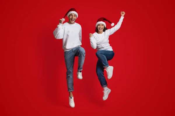 Holiday Mood. Overjoyed Couple Wearing Santa Hats Jumping On Red Studio Background — Stock Photo, Image