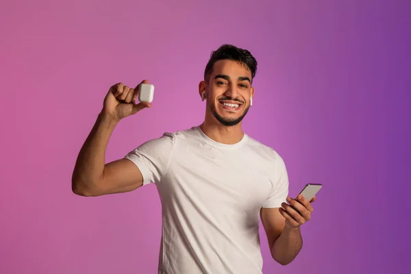 Wesoły młody Arab używa smartfona, pokazuje słuchawki bezprzewodowe, reklamuje nowy gadżet w neonowym świetle — Zdjęcie stockowe