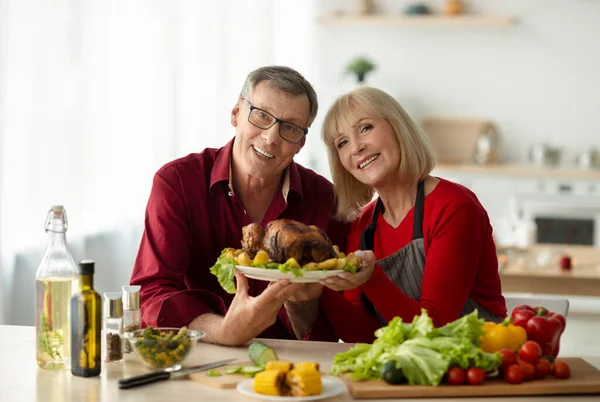 Retrato de feliz pareja de ancianos sosteniendo plato con sabroso pavo asado para la celebración familiar de Navidad en la cocina —  Fotos de Stock