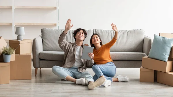 Millennial pareja asiática sentada en el suelo entre cajas de cartón, utilizando tableta digital, soñando con el diseño de un nuevo hogar —  Fotos de Stock