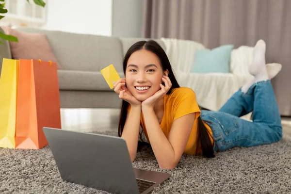 オンラインで服を注文。ラップトップとクレジットカードでカーペットの上に横たわる幸せなアジアの女性,自宅からウェブストアでのショッピング — ストック写真