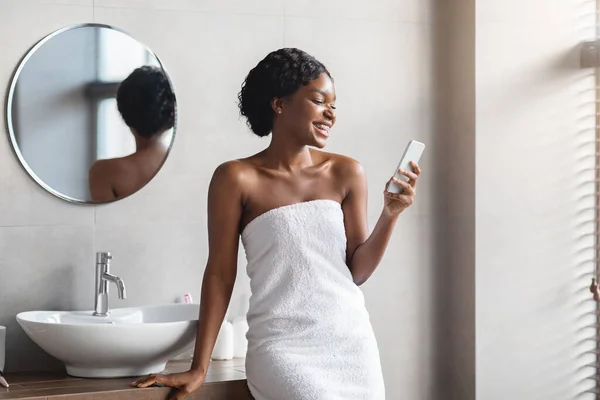 Vrolijke jonge zwarte vrouw bedekt met handdoek met behulp van smartphone — Stockfoto