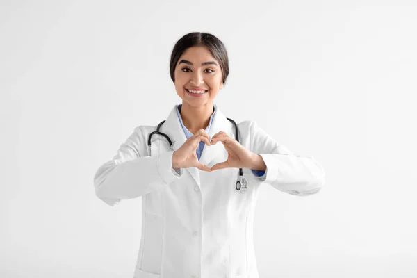 Sonriente mujer india bastante joven médico en abrigo con estetoscopio hace signo de corazón con las manos sobre fondo blanco —  Fotos de Stock