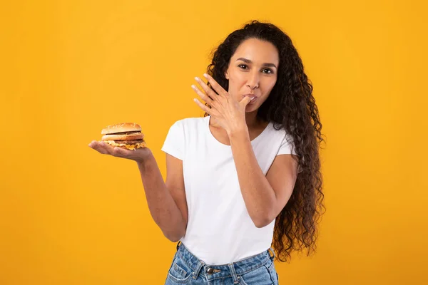 Feliz latina dama holding hamburguesa lamiendo dedo en estudio — Foto de Stock