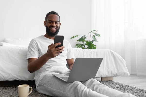 Gülümseyen, sakallı, laptopta ve telefonda konuşan, yatak odasında sohbet eden Afro-Amerikan genç adama sevindim. — Stok fotoğraf