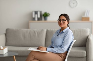 Kendine güvenen Arap kadın psikolog, panoya yazı yazıyor modern ofiste oturuyor ve kameraya gülümsüyor.