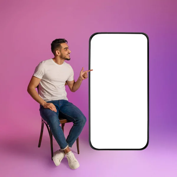 Feliz joven árabe sentado en la silla, apuntando a un teléfono celular gigante con pantalla blanca vacía en luz de neón, maqueta —  Fotos de Stock