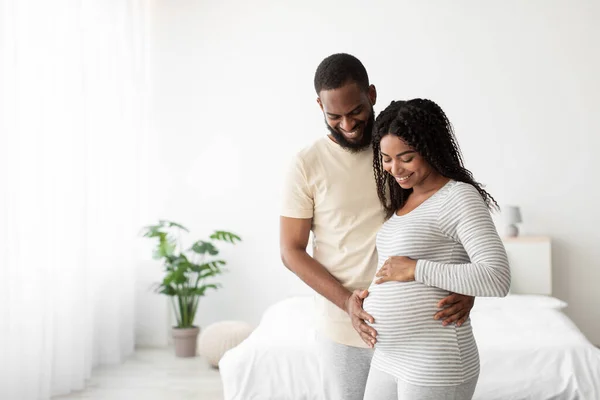 Gülümseyen Afro-Amerikan kocası hamile karısına sarılıyor, minimalist yatak odasında karnına dokunuyor. — Stok fotoğraf