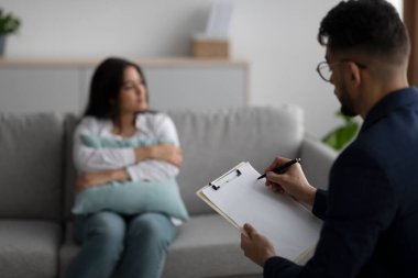 Üzgün genç Arap kadın yastığa sarılıyor ve psikologla konuşuyor, psikiyatriste odaklanıp not alıyor.