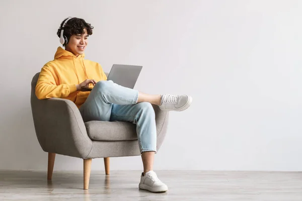 快乐的亚洲年轻人，用笔记本电脑，头戴耳机，靠着白墙坐在扶手椅上，复制空间 — 图库照片
