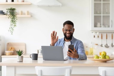 Gülümseyen genç Afro-Amerikan sakallı adam dizüstü bilgisayarla telefonda web kamerasına el sallıyor.