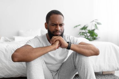 Düşünceli, üzgün, beyaz elbiseli, siyah sakallı bir adam yerde, yatağın yanında oturuyor.