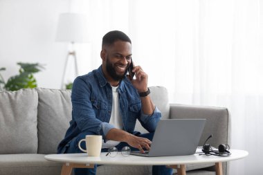 Mutlu Milenyum Afro-Amerikan erkekleri dizüstü bilgisayarda çalışır ve iş yerinde oturma odasındaki koltukta telefonla konuşur.