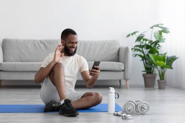 Gülümseyen siyah sakallı bir sporcu yere oturmuş dambıllar ve su şişesiyle el sallıyor.