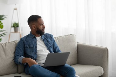 Ciddi şekilde konsantre olmuş siyah sakallı bir adam. İç odadaki kanepede dizüstü bilgisayarda çalışıyor. Pencereye bak.