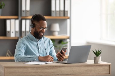 Gözlüklü ve kulaklıklı neşeli Afro-Amerikan sakallı adam laptopta çalışıyor ve akıllı telefondan sohbet ediyor.