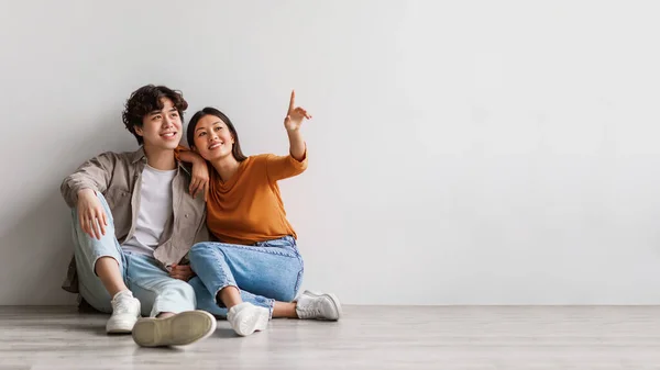 Carefree millennial Asiático homem e mulher sentado no chão perto da parede do estúdio branco e apontando para o espaço de cópia, design de banner — Fotografia de Stock