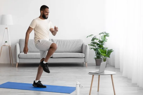 Χαμογελαστός νεαρός μαύρος αθλητής με άσπρα ρούχα κάνει ασκήσεις ποδιών, προθέρμανση, παρακολουθήστε βίντεο στον υπολογιστή στο εσωτερικό του δωματίου — Φωτογραφία Αρχείου