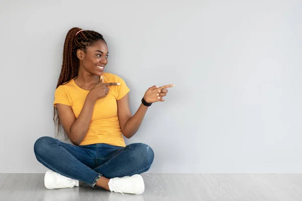 Классное предложение. Счастливая молодая черная женщина, сидящая скрестив ноги на полу, указывая в сторону копирования пространство против серой стены студии — стоковое фото