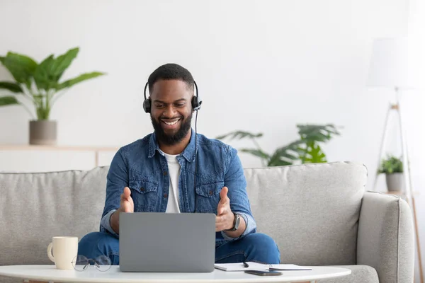 Blij vriendelijke jonge Afrikaanse Amerikaanse bebaarde zakenman in headset werk op de computer op de bank — Stockfoto