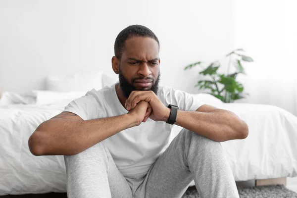 Pensativo molesto joven negro barbudo hombre en ropa blanca se sienta en el suelo, cerca de la cama, reflexiona — Foto de Stock