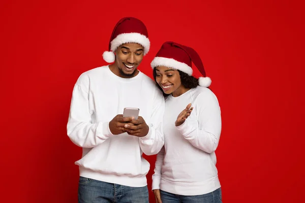 Cheerful black couple in Santa hats using smartphone — Stock Photo, Image