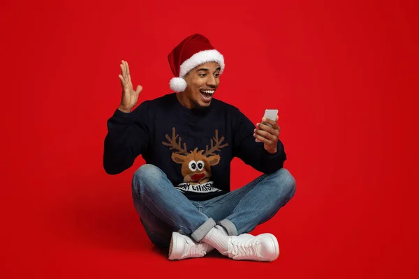 Noel Baba şapkalı mutlu Afrikalı Amerikalı adam akıllı telefon kullanıyor, el kol hareketi yapıyor. — Stok fotoğraf