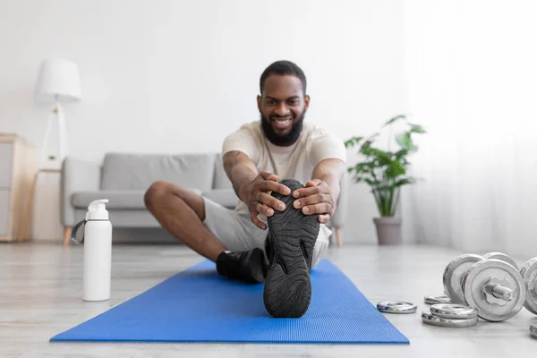 Happy millennial black bearded sportsman make exercise for legs, stretching on floor on mat with dumbbells — 图库照片