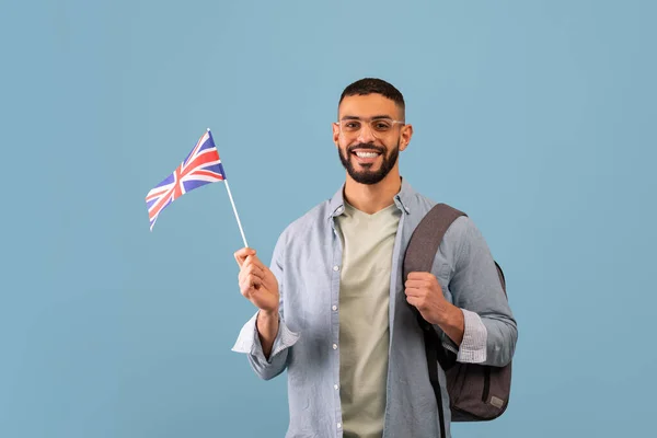 Happy arab guy with backpack showing flag of Great Britain over blue studio background, male student studující angličtinu — Stock fotografie
