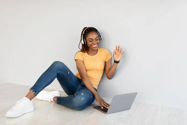 Fiatal fekete hölgy használja laptop pc az online kommunikáció, ül a padlón, integetett webkamera ellen szürke stúdió fal — Stock Fotó